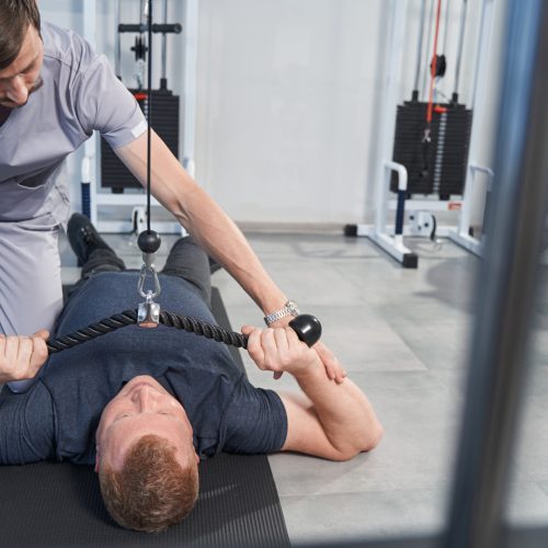 Patient exercising on tether machine with physiotherapist in rehabilitation gym. Man with disability