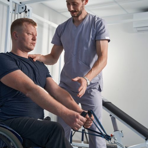 Wheelchair young man stretch elastic strap on weight training machine in rehab clinic with doctor